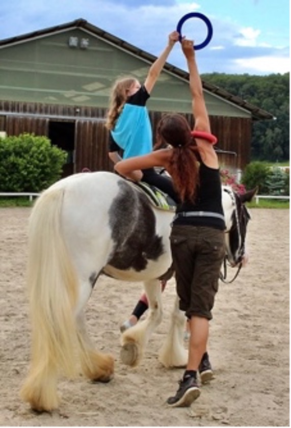 Ein Bild das die Hippotherapie zeigt. Zu sehen ist das Pferd, ein Mädchen sitzt darauf und die Therapeutin daneben hält einen Ring hoch, den das Mädchen mit gestrecktem Arm ergreift.

          